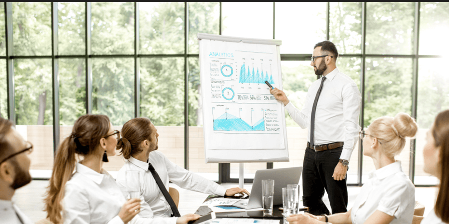 Un bărbat prezintă date "Analytics" pe un flip chart către o echipă într-o sală de ședințe luminoasă.