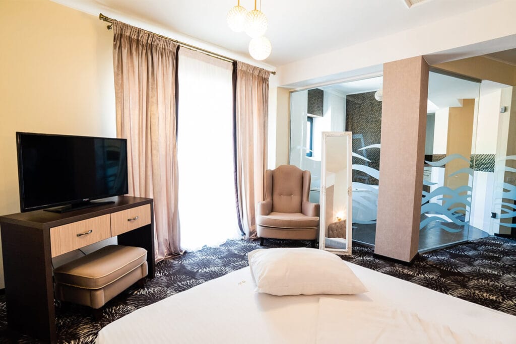 Interiorul camerei de hotel cu TV, fotoliu, oglindă înaltă și fereastră cu perdele.