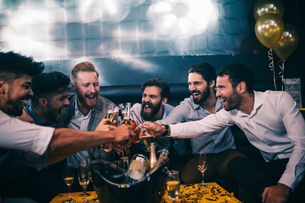 Un grup de prieteni ciocnesc sticle de bere, zâmbitori, într-un bar cu baloane aurii.