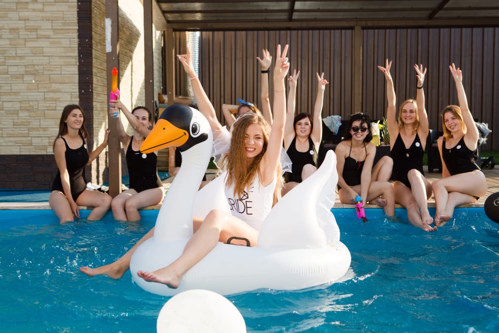 Un grup de femei se bucură de o petrecere a burlăcițelor într-o piscină cu o mireasă pe o lebădă gonflabilă.