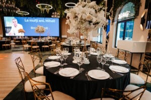 Sărbătoarea "La mulți ani, Mihail" într-un restaurant cu mese rotunde negre, flori albe, scaune aurii și un ecran mare.