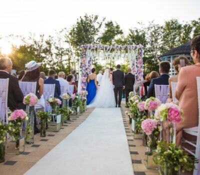 O nuntă în aer liber, cu invitați așezați pe scaune decorate, cu un culoar alb și un cuplu în fața altarului floral.