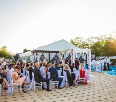 O ceremonie de nuntă în aer liber, cu o piscină și invitați așezați pe scaune albe.