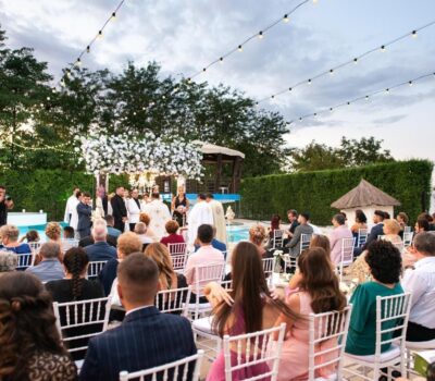 Căsătorie în aer liber. Mulțimea este așezată pe scaune albe. O piscină și verdeață bogată în fundal.