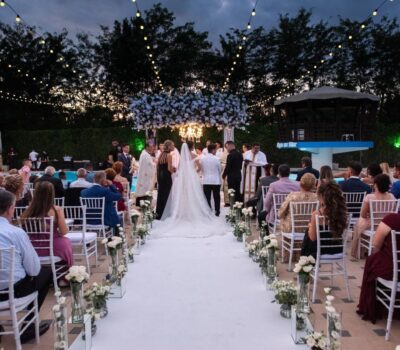 Ceremonia de nuntă în aer liber cu invitați așezați, mireasă și mire pe culoar, aranjament floral și lumini deasupra.