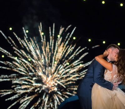 Un cuplu se sărută pe fundalul unui foc de artificii, luminile șirului deasupra. Mireasa poartă o rochie albă. Mirele poartă un costum bleumarin.