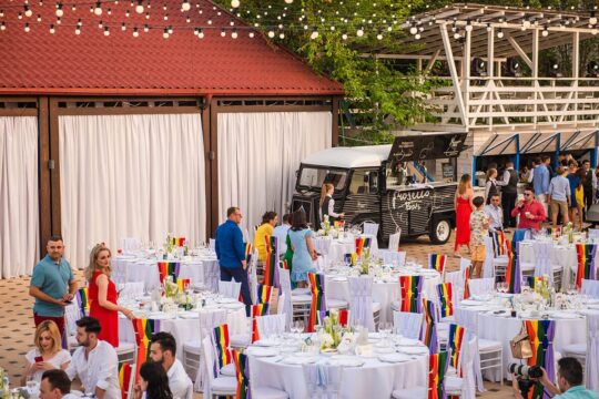 O nuntă cu mese albe, scaune decorate în curcubeu, un bar Prosecco și șiruri luminoase deasupra.