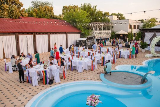 Nuntă în aer liber cu mese rotunde, piscina și mulți invitați.