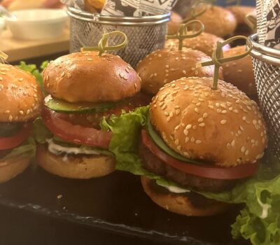 Mini burgeri cu susan, roșii, castraveți și salată, pe o farfurie neagră.