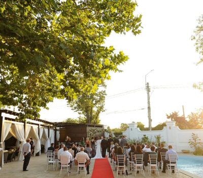 Nunta în aer liber, cu oaspeți așezați de-a lungul unui culoar roșu care duce la mireasă și mire.