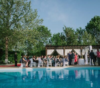 Nuntă în aer liber lângă piscină, cu invitați așezați și preot cu veșminte roșii.