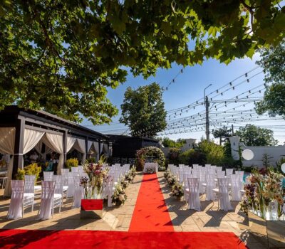 Aranjament de nuntă în aer liber cu covor roșu, flori și scaune albe.