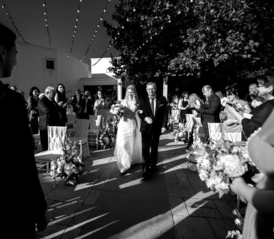 Fotografie alb-negru cu o mireasă condusă pe culoar de tatăl ei, cu invitați pe ambele părți.