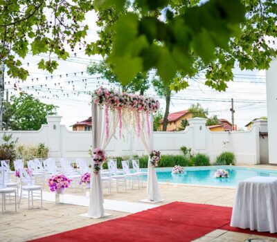 Scena nunții în aer liber cu aranjament floral, rânduri de scaune albe și piscină.