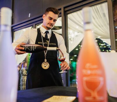 Un somelier de la Wine Taster School toarnă vin roșu într-un pahar. Sticle de vin albe și roz sunt în prim-plan.