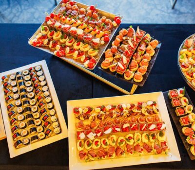 Diverse aperitive aranjate pe platouri pe o masă neagră, inclusiv sushi și canapés.