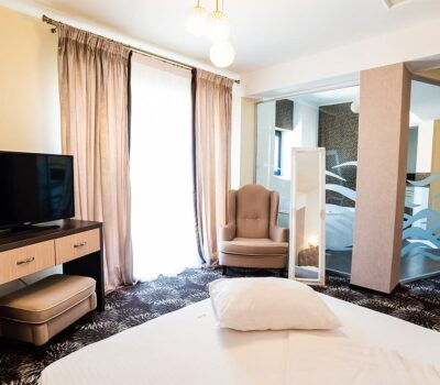 Interiorul unui dormitor luminos, cu un televizor, un scaun și o baie parțial vizibilă.