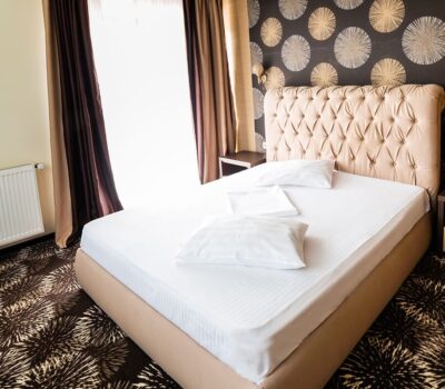 Camera de hotel cu pat dublu, lenjerie albă, tăblie tapițată și noptiere. Tapet cu model circular în fundal.