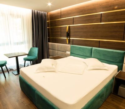 Interior de cameră de hotel modernă cu pat verde-albastru, noptiere și scaune lângă fereastră. Panouri decorative din lemn pe perete.