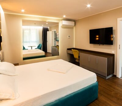 Interiorul unei camere de hotel, cu pat, oglindă, birou și televizor.