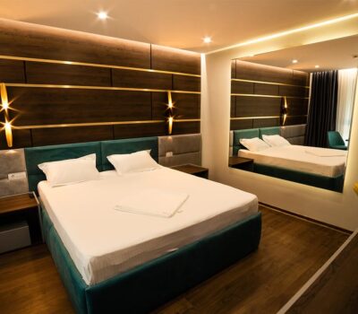 Dormitor cu pat dublu, lenjerie albă, tăblie verde/gri. Un perete are panouri din lemn și o oglindă mare.