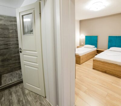 Interior cu baie parțial deschisă și dormitor cu două paturi de o persoană și tăblii turcoaz.