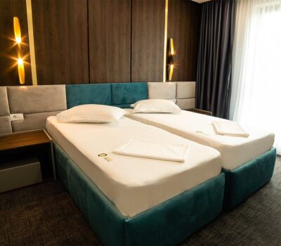 Interior cu două paturi de o persoană, tăblii tapițate și noptieră, iluminat decorativ și draperii.