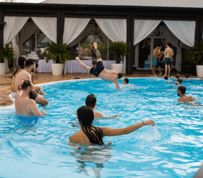 Oameni într-o piscină în aer liber, cu un bărbat care face o săritură în aer.