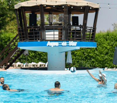 Oameni se joacă cu o minge în piscină, cu o structură „Aqua-del-Mar” în fundal.