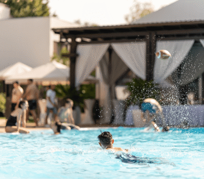 Oamenii înoată și se joacă într-o piscină în aer liber. Un fotbal aerian poate fi văzut în fundal.