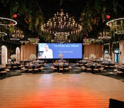 O sală de evenimente decorată cu candelabre, plante și textul "La mulți ani, Mihail! Bine ați venit!".