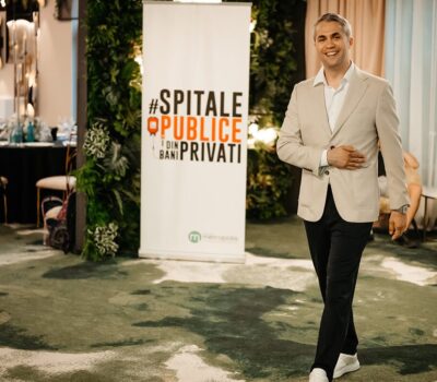 Un bărbat zâmbitor, îmbrăcat la patru ace, lângă un banner "#spitale publice din bani privați".