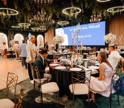 Interior decorat festiv cu "La mulți ani, Mihai!" pe ecran. Oameni așezați la mese festive.