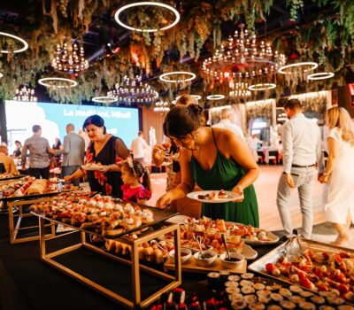 Oamenii servesc mâncare la un eveniment cu textul „La Mulți Ani, Mihai” pe un ecran.