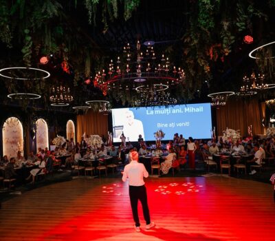 Interiorul unui eveniment cu o mulțime de oaspeți. Un ecran mare spune: "La mulți ani, Mihail! Bine ați venit!"