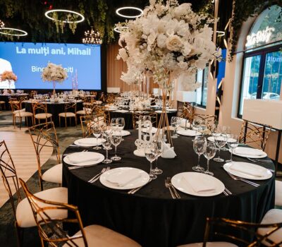 Mese rotunde cu fețe de masă negre, aranjamente florale albe. Ecranul prezintă "La mulți ani, Mihail!"