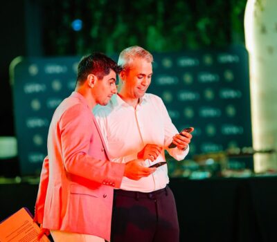 Doi bărbați se uită la telefoanele mobile. Unul poartă un sacou roz, iar celălalt o cămașă albă.