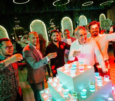 Bărbați care ciocnesc shot-uri. O piramidă de shot-uri colorate pe o platformă iluminată. Interior decorat cu verdeață.