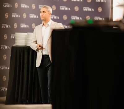 Un bărbat în costum bej stă în fața unui fundal "SAUTICA".