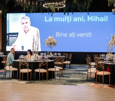 Interior cu mese pentru o petrecere. Pe un ecran scrie: "La mulți ani, Mihai! Bine ați venit!".