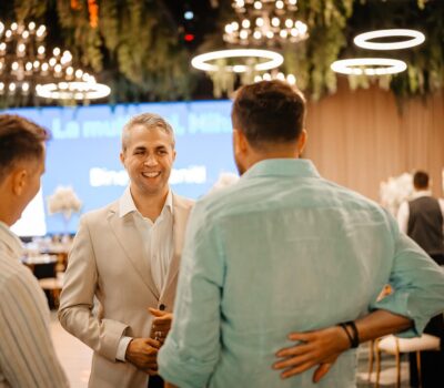 Trei bărbați într-o cameră; unul poartă un costum bej și zâmbește. În fundal apare text.