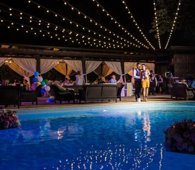 Eveniment festiv de noapte lângă piscină, cu luminițe, baldachin și oaspeți îmbrăcați elegant.