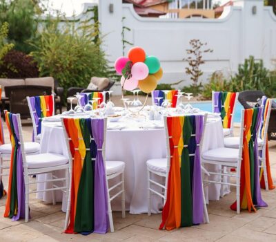 Masa rotundă cu scaune albe decorate cu curele curcubeu și baloane.