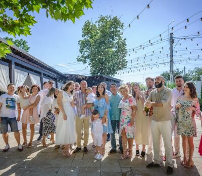 Un grup de oameni sărbătoresc cu șampanie într-o zi însorită, cu lumini suspendate.