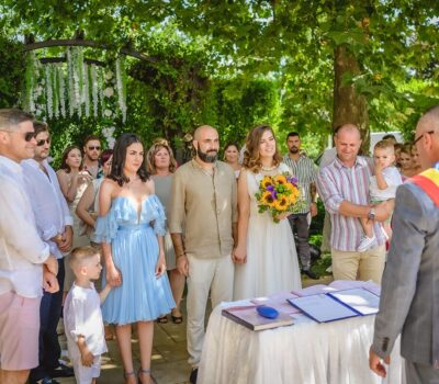 Ceremonie de nuntă în aer liber, cu mireasă cu floarea soarelui, mire și oficial cu eșarfă tricoloră. Oaspeți prezenți.