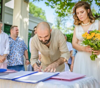 Un bărbat cu barbă semnează documente lângă o mireasă cu un buchet de flori, martori privind.