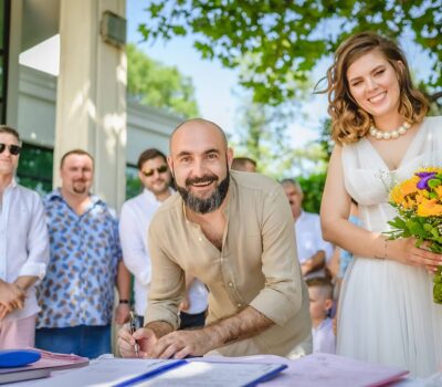 Un bărbat semnează un document nupțial cu o mireasă zâmbitoare și invitați în fundal.