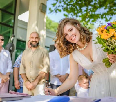 Mireasă zâmbitoare în rochie albă, cu buchet, semnând un document.