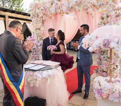 Un cuplu se căsătorește sub un arc floral. Un ofițer de stare civilă poartă o eșarfă românească. Invitatii aplaudă.