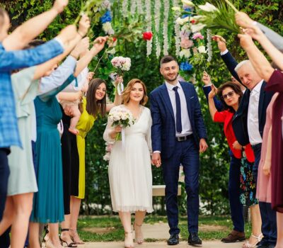 Un cuplu căsătorit merge printr-o mulțime de oameni cu flori ținute sus în aer, în timpul unei nunți în aer liber.